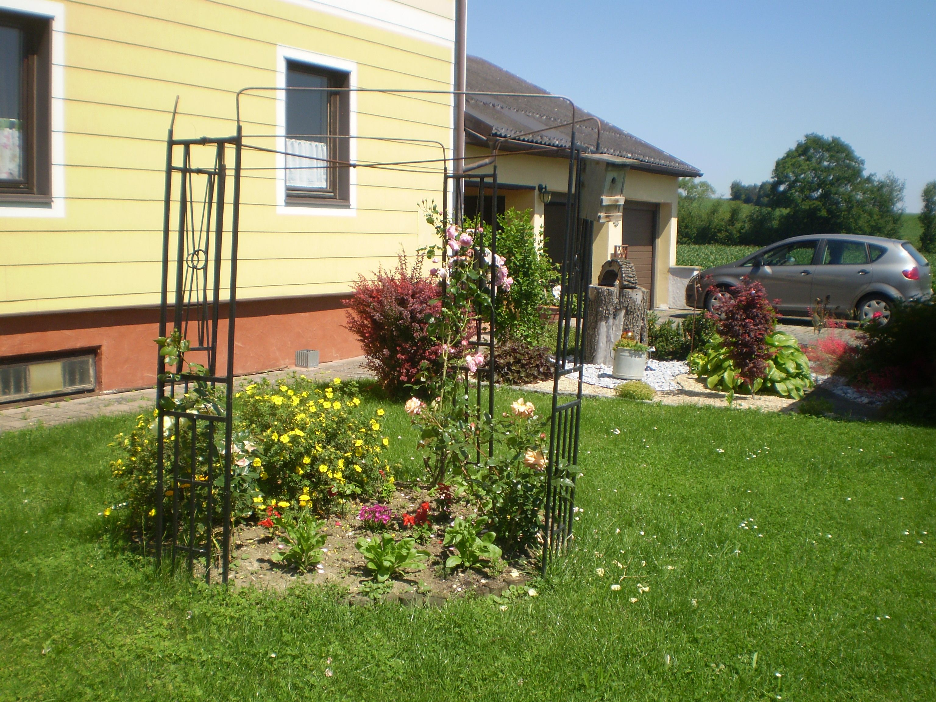 A well-tended garden with a flower bed and trellis in front of a yellow house.