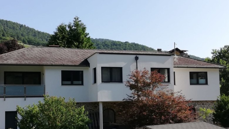 Exterior view of vacation home Johanna, © Familie Schoßmann Exterior view of a modern white vacation home with trees in the foreground and wooded hills in the background.