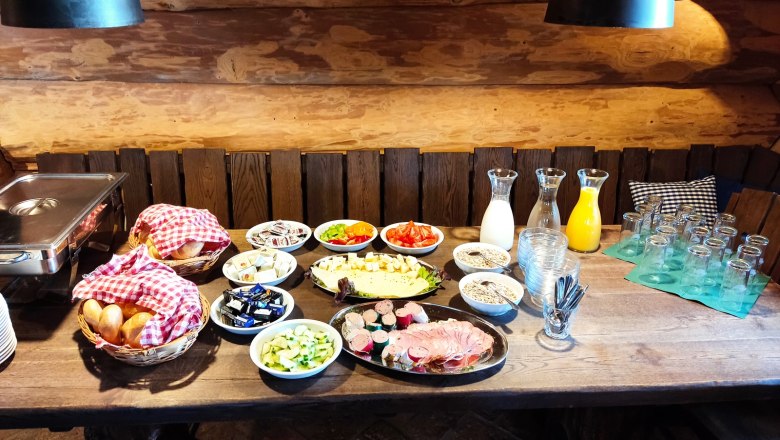 Breakfast buffet with bread rolls, cold cuts, cheese, vegetables, muesli and drinks on a wooden table.