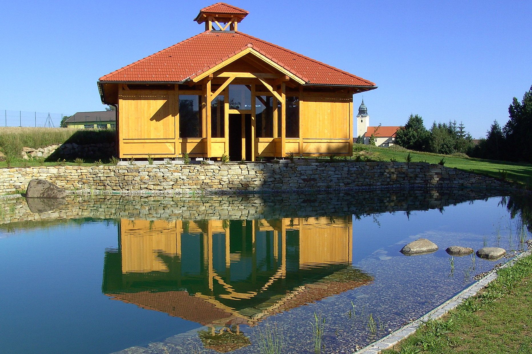 A wooden building with a red roof is reflected in a pond, surrounded by a green landscape and blue sky.