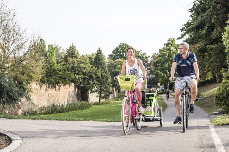 A couple is riding bicycles on a park path. The woman is pulling a child's trailer.