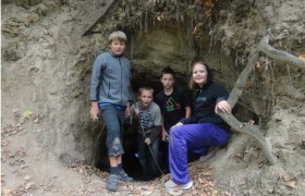 Four children are standing in front of a cave entrance in a forest.
