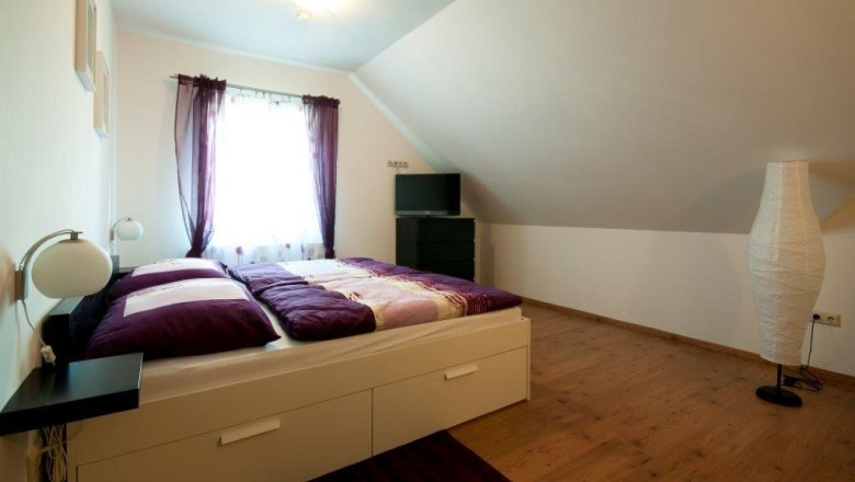 A cozy guest room with a double bed, purple curtains and a TV on a chest of drawers under a sloping roof.