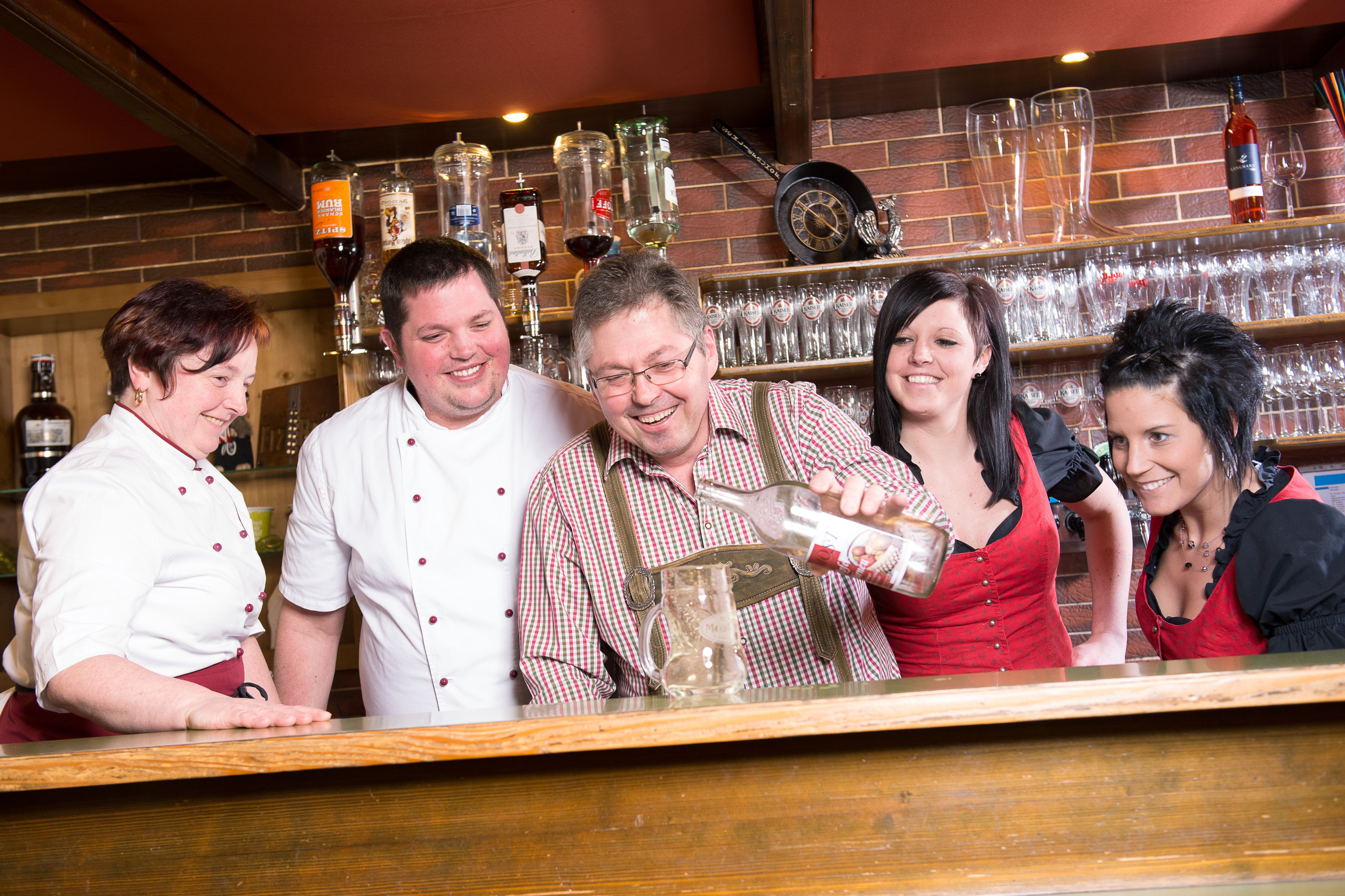 A group of people behind a bar, laughing together and pouring a bottle into a jug.