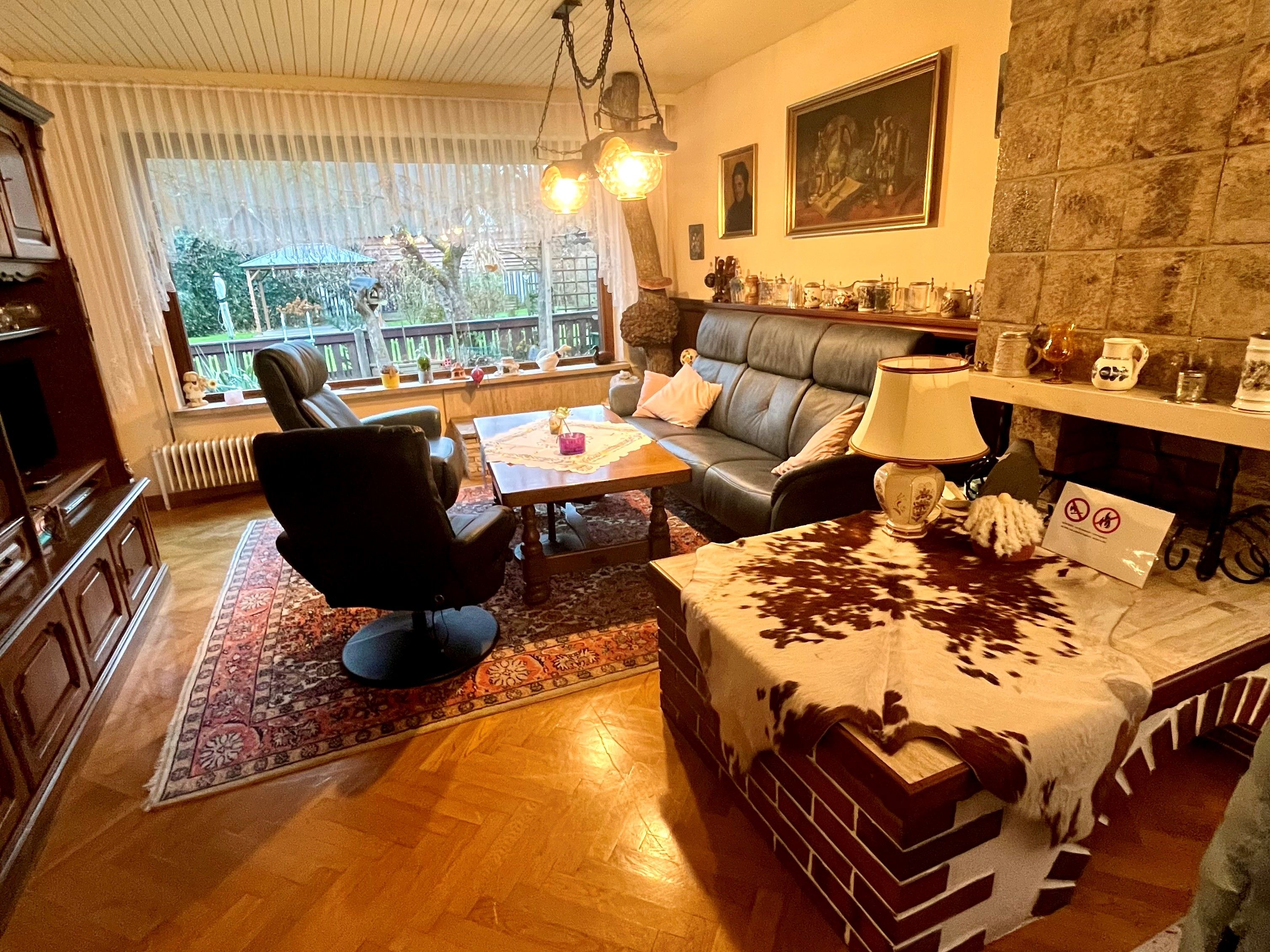 Cozy living room with leather sofa, armchair, wooden table and fireplace. Large window front with view to the outside. Rustic furnishings with carpet.