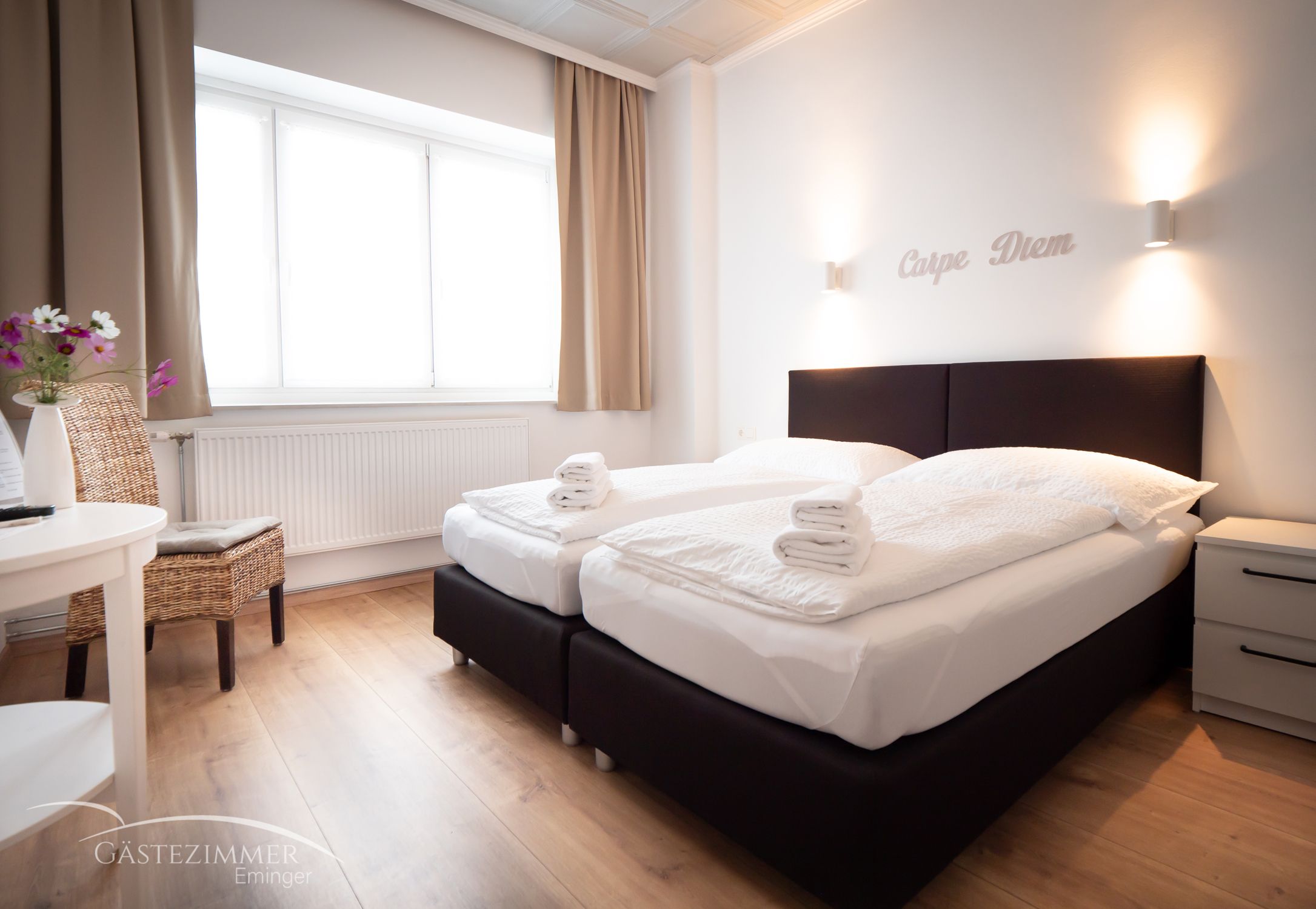 Modern double room with two beds, white comforters and towels. 'Carpe Diem' lettering on the wall, light-colored curtains and wooden floor.