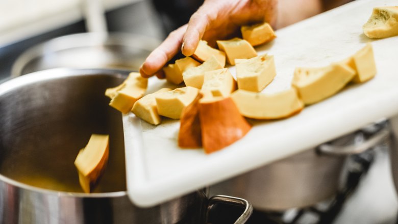 A hand pushes sliced pumpkin pieces from a cutting board into a pot.