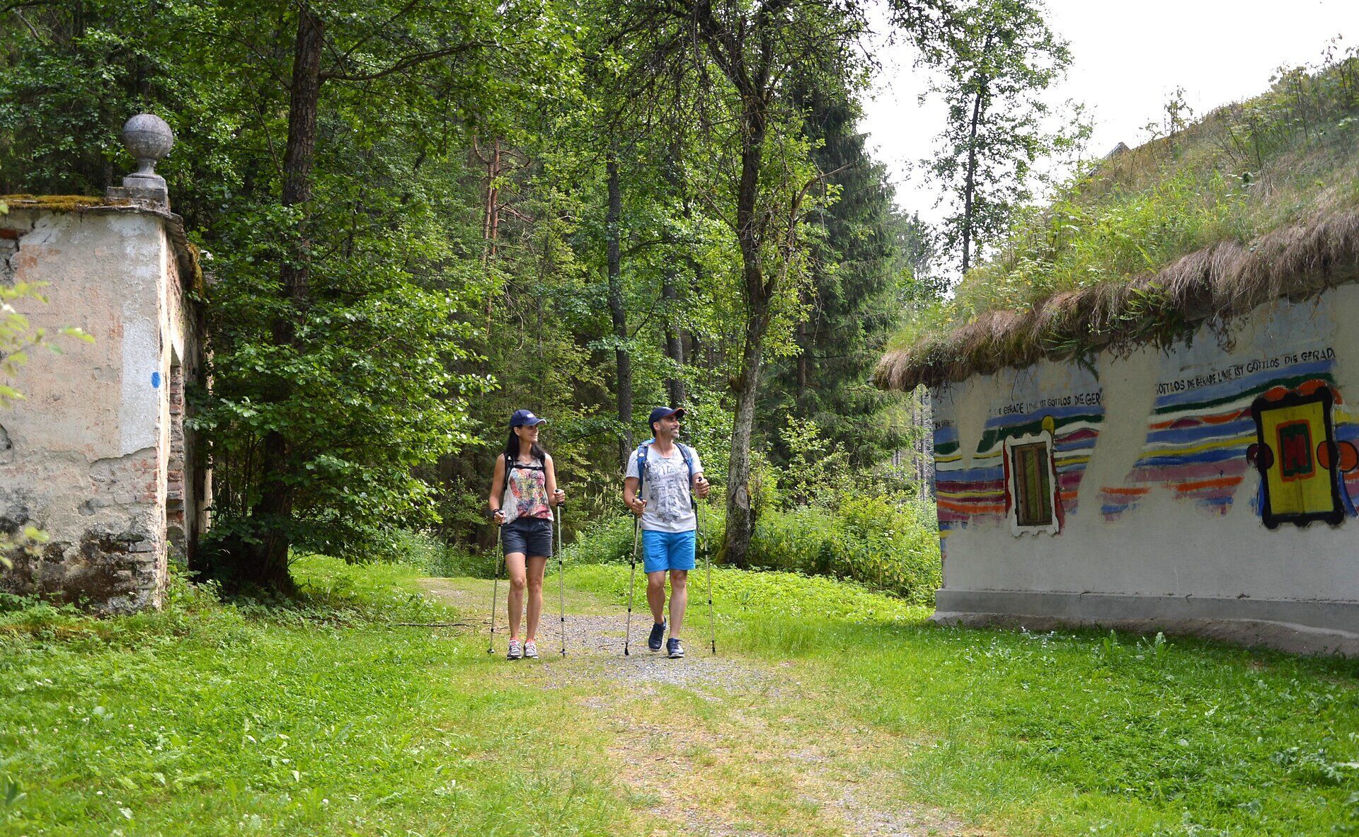 Ein Paar wandert entspannt auf einem schmalen Pfad, umgeben von üppigem Grün und der beruhigenden Stille des Waldes. Die bunten, kunstvoll gestalteten Wände der Hundertwassermühle verleihen der Umgebung einen einzigartigen Charme und laden dazu ein, die kreative Verbindung von Natur und Kunst zu entdecken.