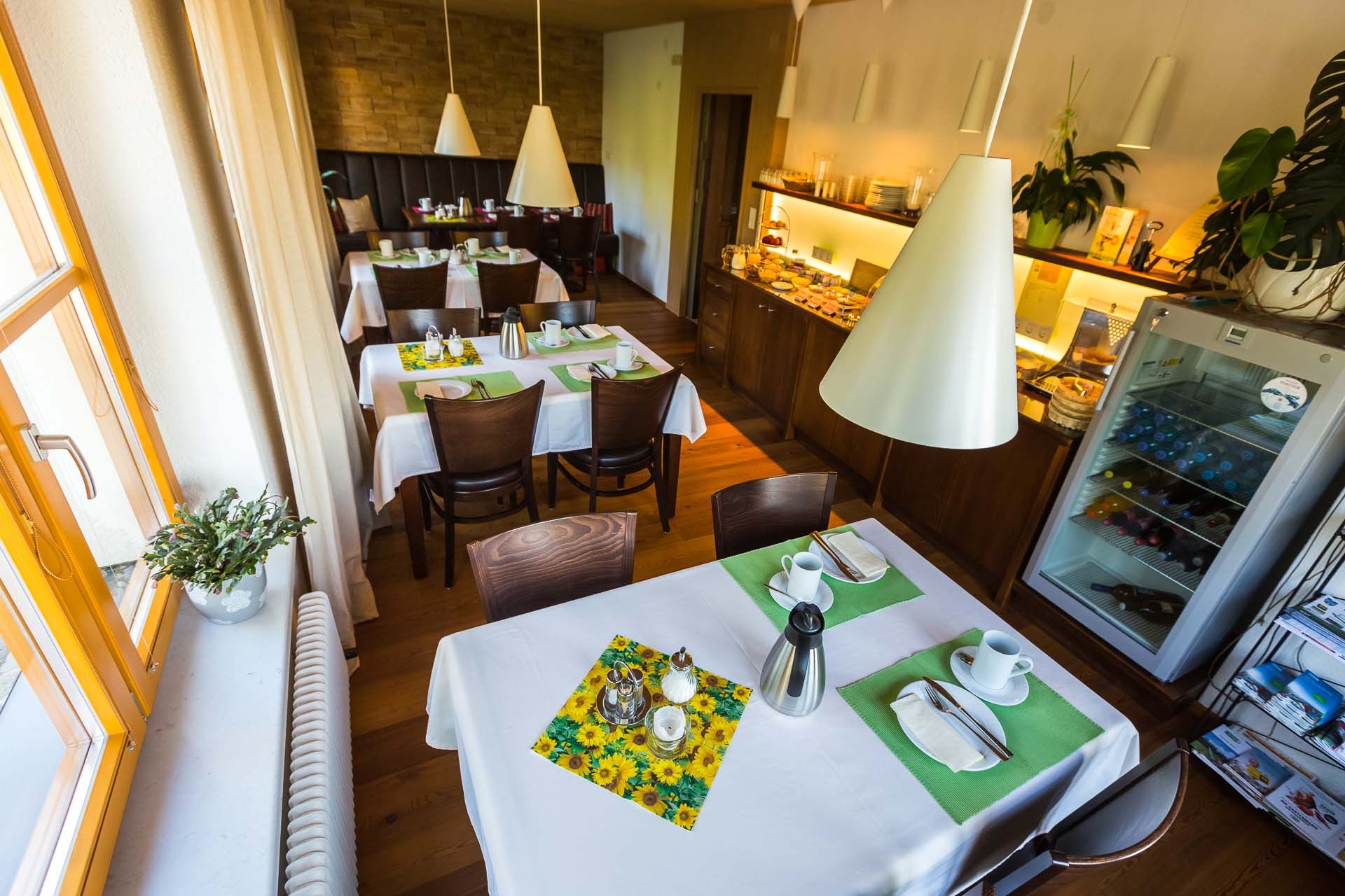 A cozy breakfast room with laid tables, white tablecloths and green placemats. A buffet and a fridge in the background.