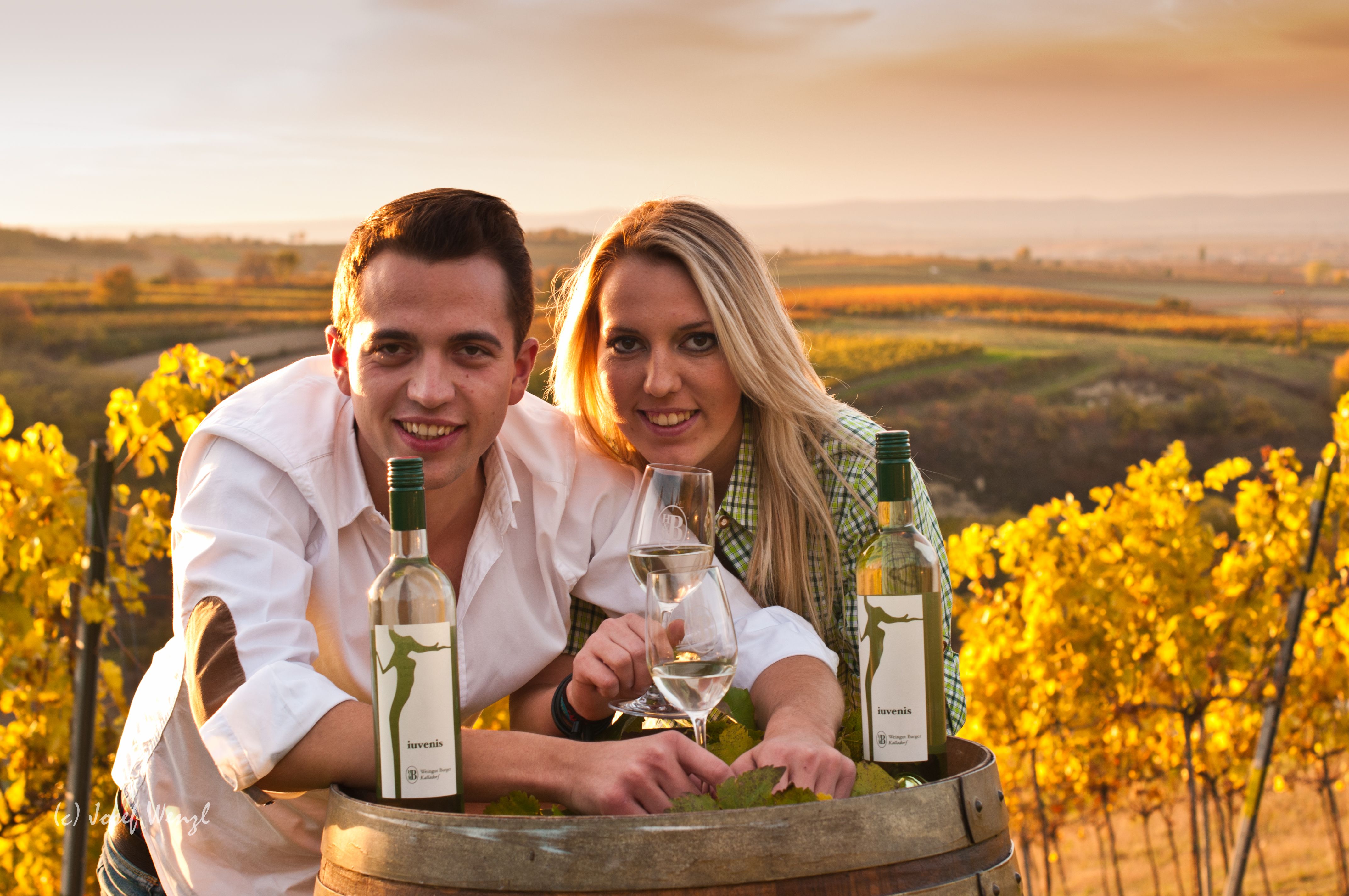 Two people lean over a wine barrel in a vineyard at sunset.