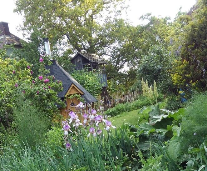 An idyllic garden with a small wooden house, surrounded by flowering plants and trees.