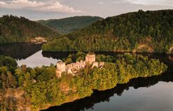 Aerial view of a castle ruin by the lake, surrounded by forest and hills.