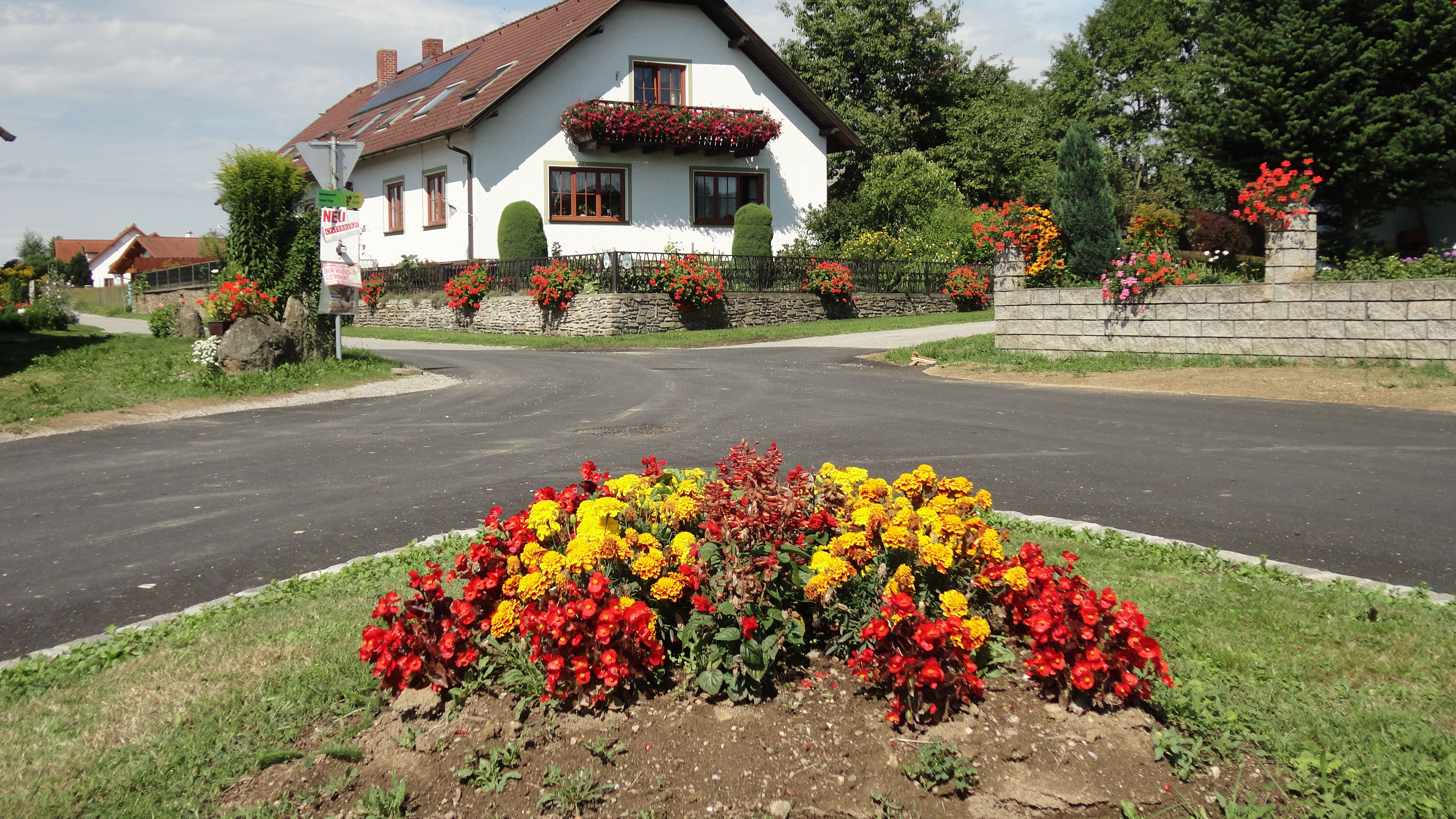 A white house with red roof tiles and floral decorations, surrounded by colorful flowerbeds and a green landscape.