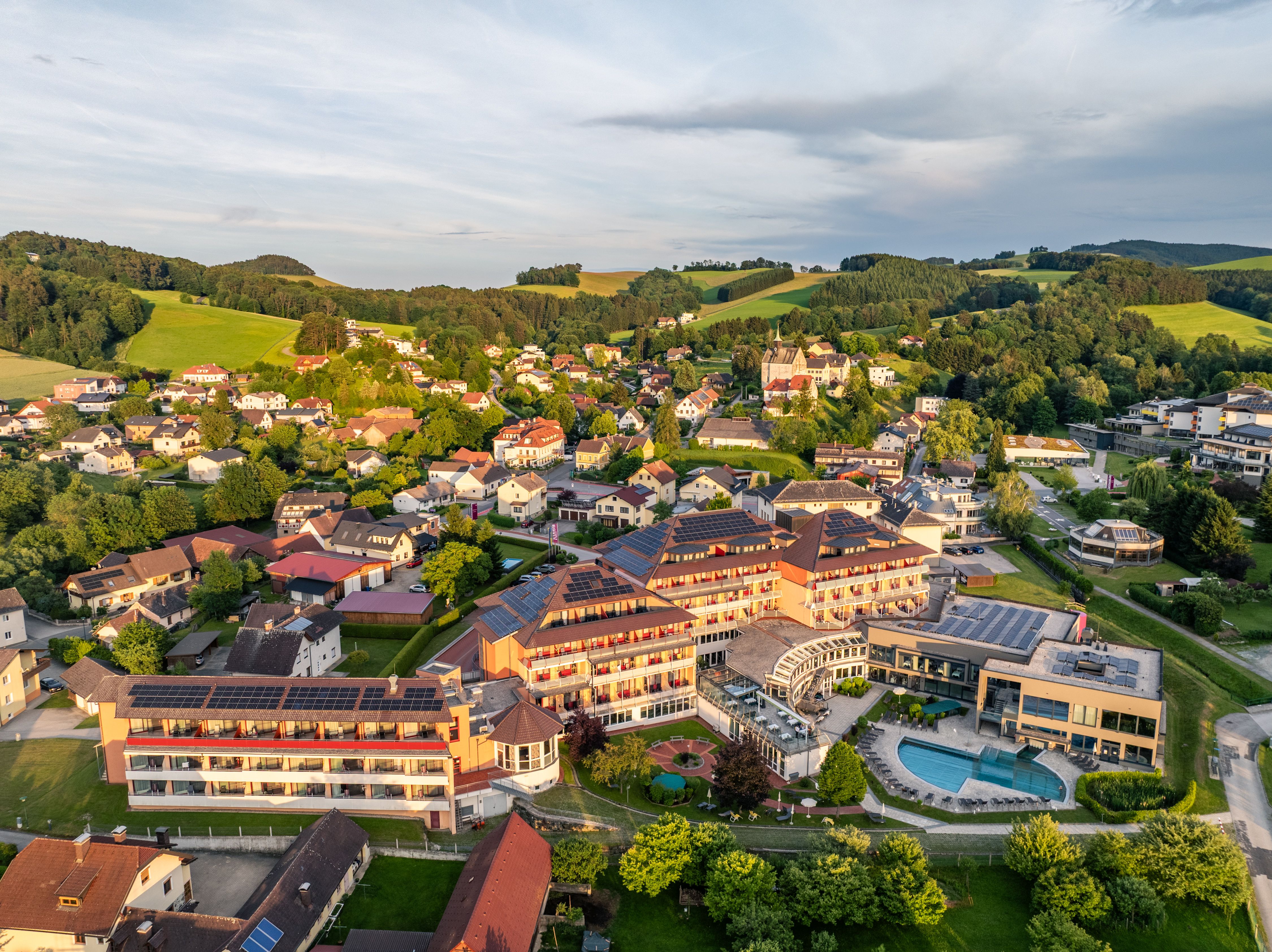 Aerial view of Bad Schönau, with a large hotel and pool in the foreground and green hills in the background.