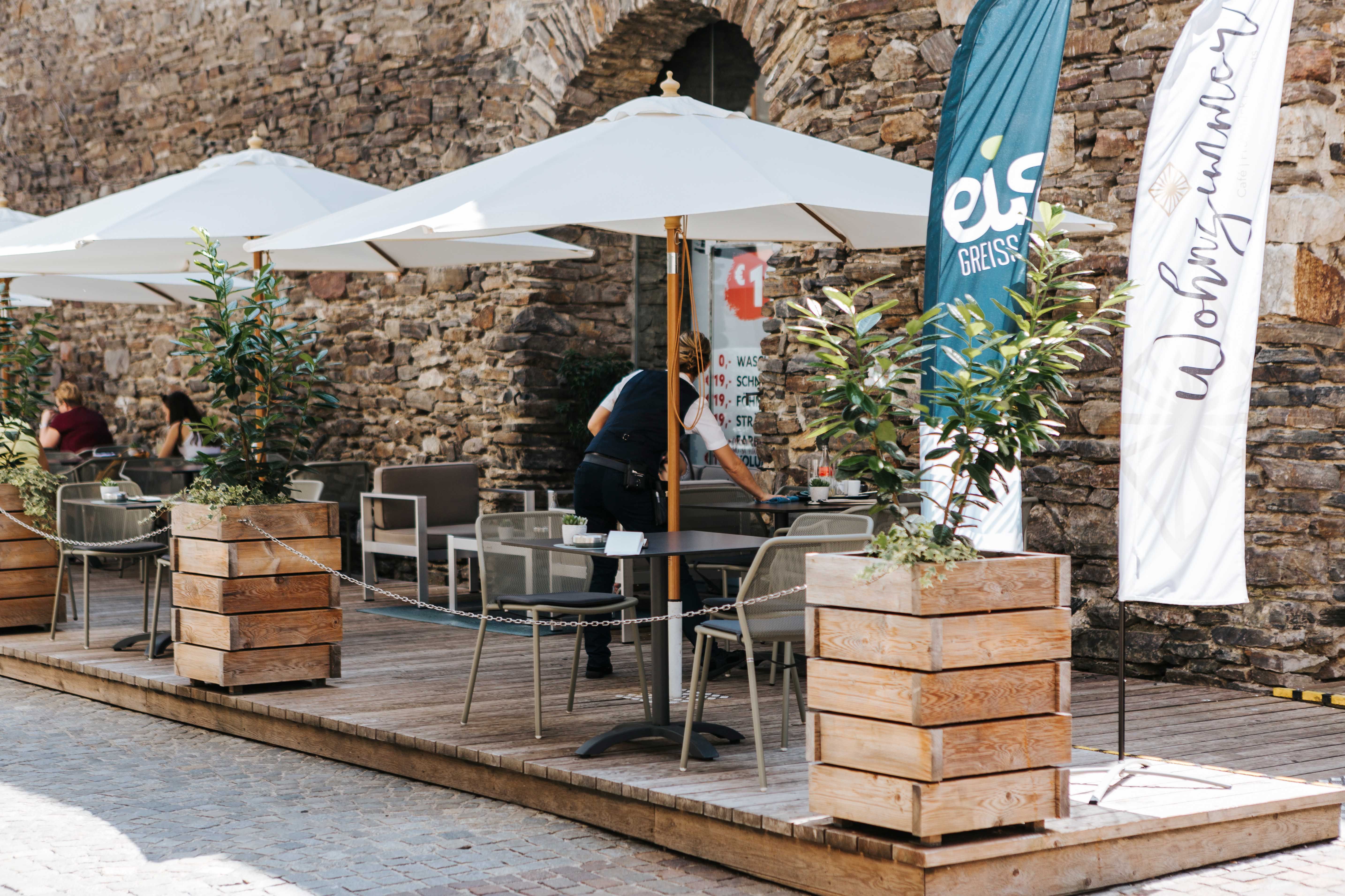 Outdoor area of a café with parasols and wooden crates as planters in front of a stone wall.