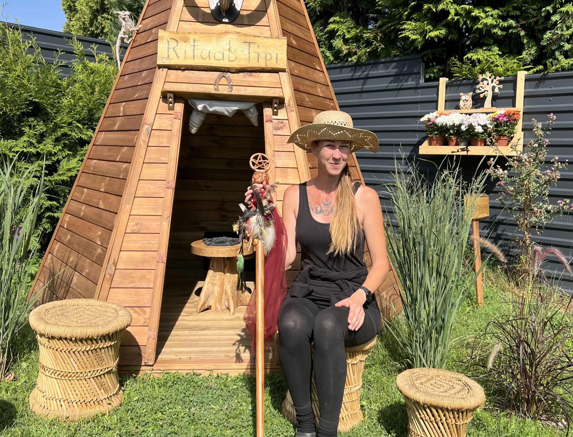 Woman in a straw hat sits in front of a wooden tepee in the garden.