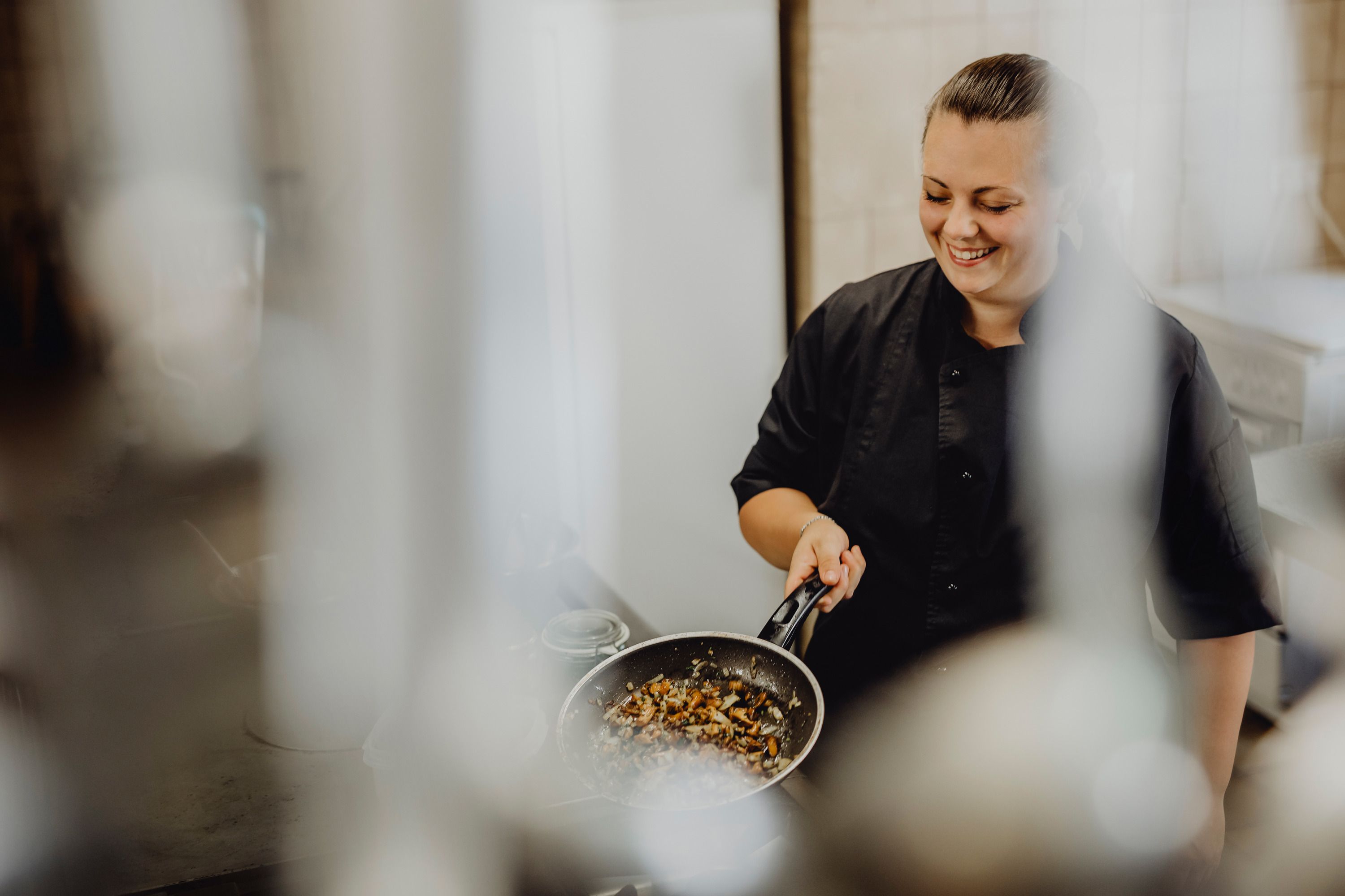 A cook in a black uniform holds a pan of roasted vegetables and smiles.