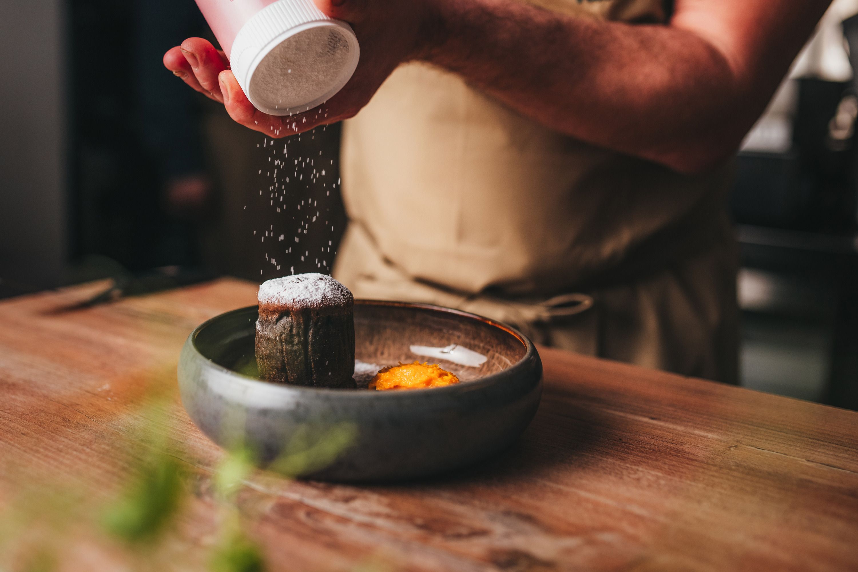 Person sprinkles chocolate cake with powdered sugar, next to it a mandarin on a plate.