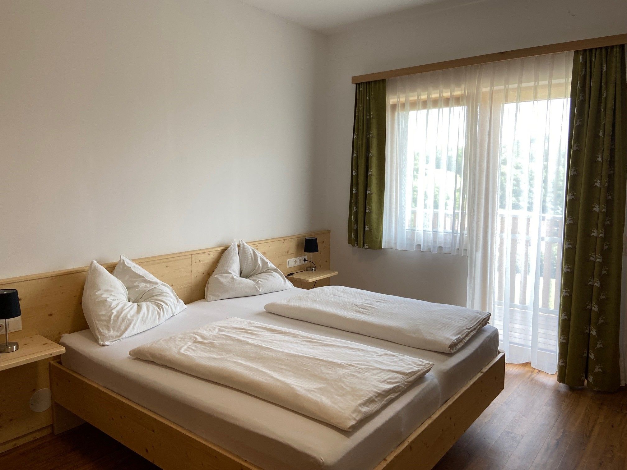 A bright bedroom with a double bed, white comforters and pillows. A window with green curtains lets in natural light.