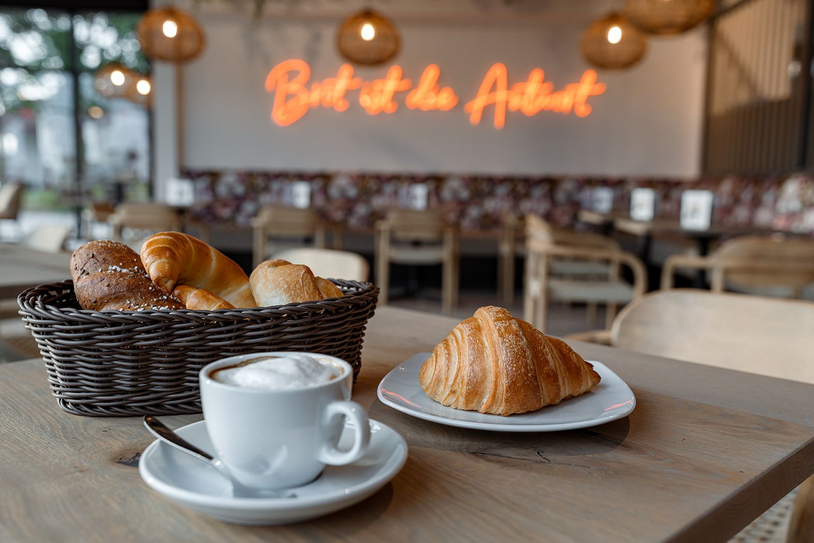 A café with a basket of pastries, a croissant on a plate and a cup of cappuccino on a table.