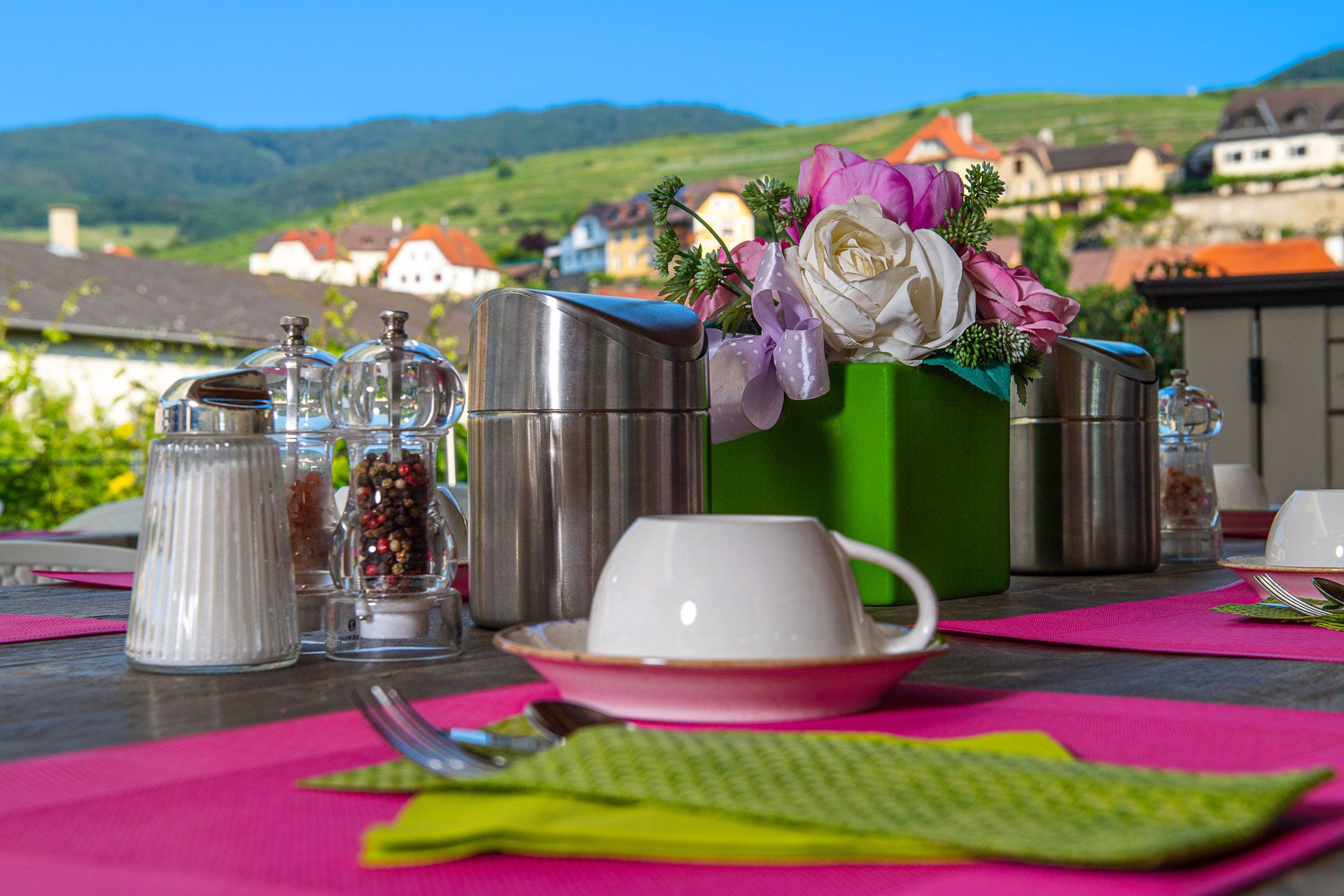 Outdoor breakfast table with pink placemats, coffee cups, salt and pepper shakers and flower arrangement, with a picturesque landscape in the background.