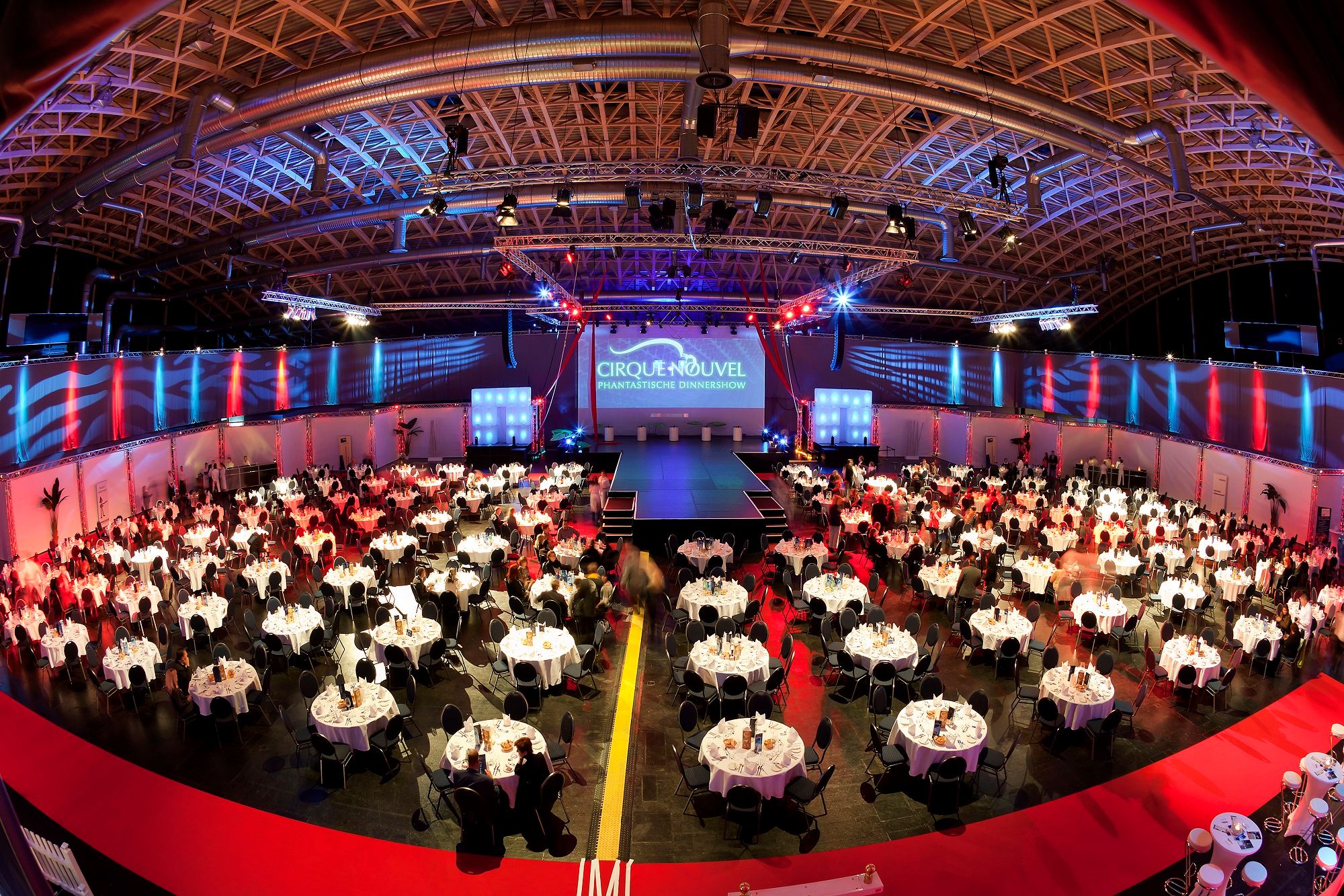 Large event room with round tables and stage, illuminated in red and blue.