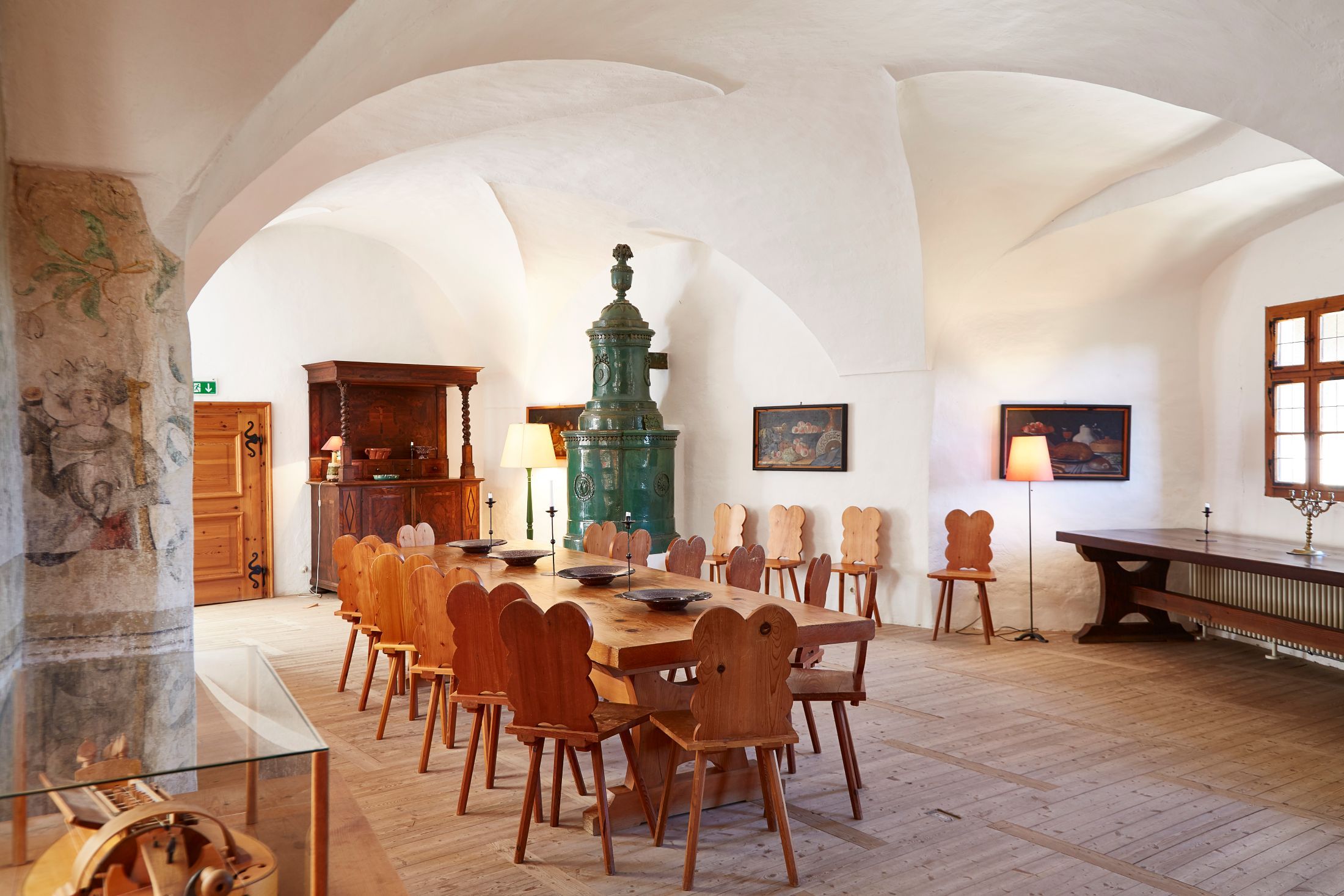 Interior view of a historic room with wooden table and chairs, a green tiled stove and wall paintings.