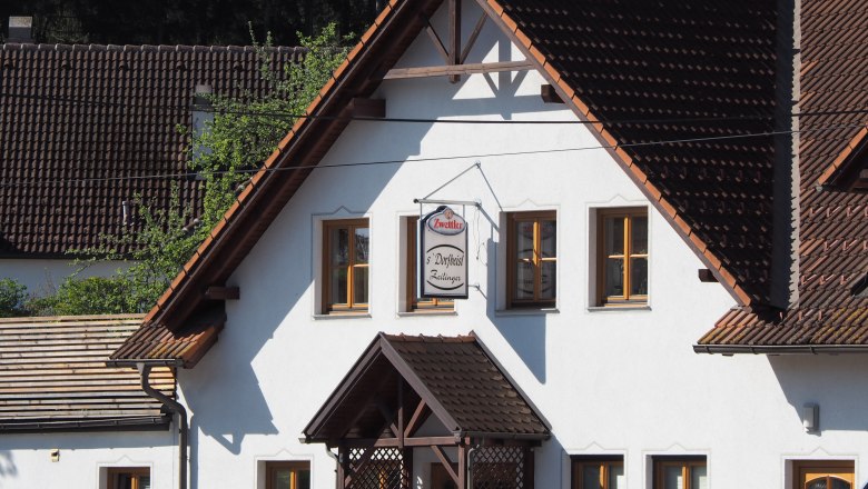 A traditional building with white walls and brown roof tiles. A sign with the inscription 'Zwettler Dorfwirt Zeilinger' hangs on the façade.