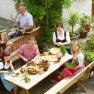 A family in traditional dress sits at a table in the garden and prepares a meal.