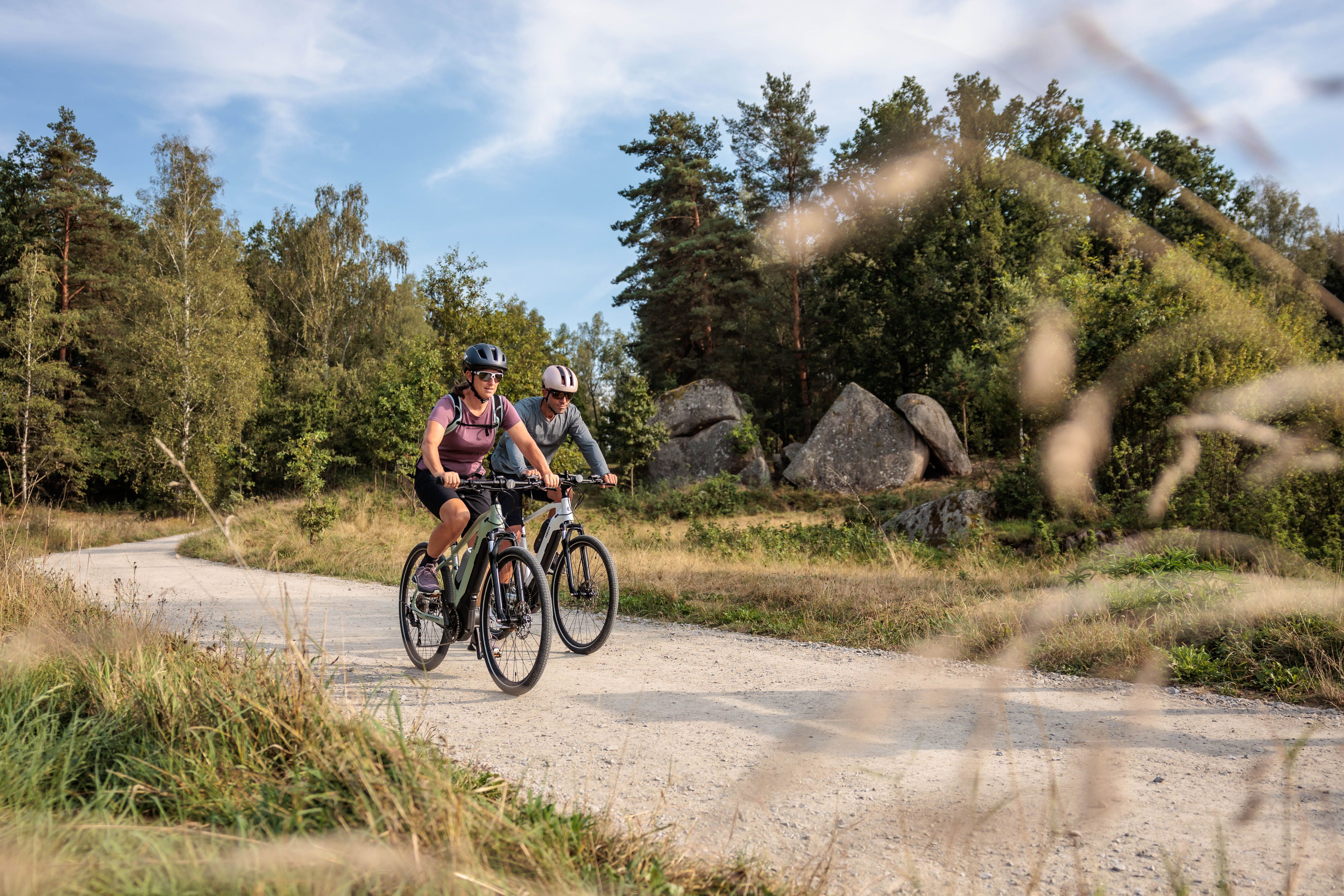 Two cyclists on a gravel path in the Blockheide Nature Park, surrounded by trees and rocks.