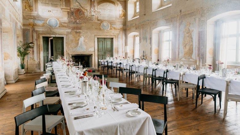 Elegant ballroom with long, laid tables, antique wall paintings and chandeliers.