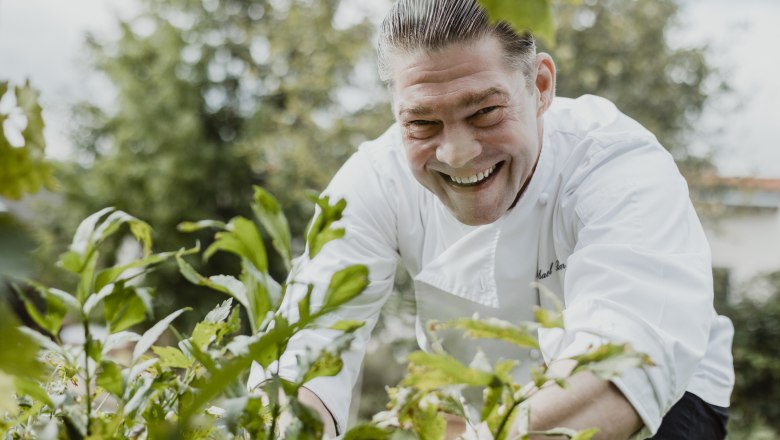 Smiling chef in white uniform in the garden, surrounded by plants.