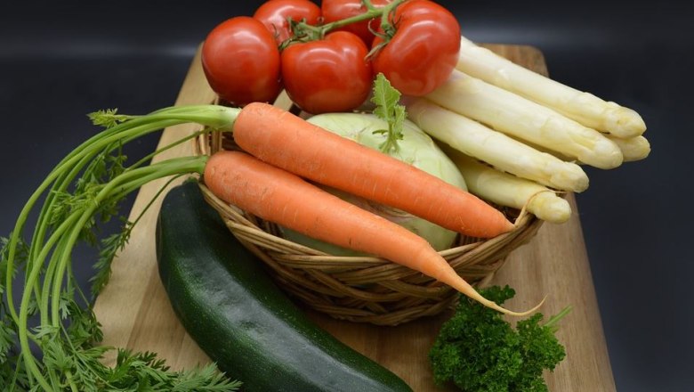 Basket of fresh vegetables on a wooden board: carrots, tomatoes, white asparagus, zucchini, cabbage and herbs against a dark background.