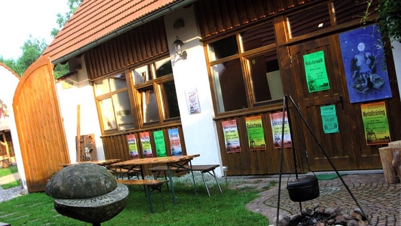 A courtyard with wooden tables, posters on a wooden wall and a cauldron over a fireplace.