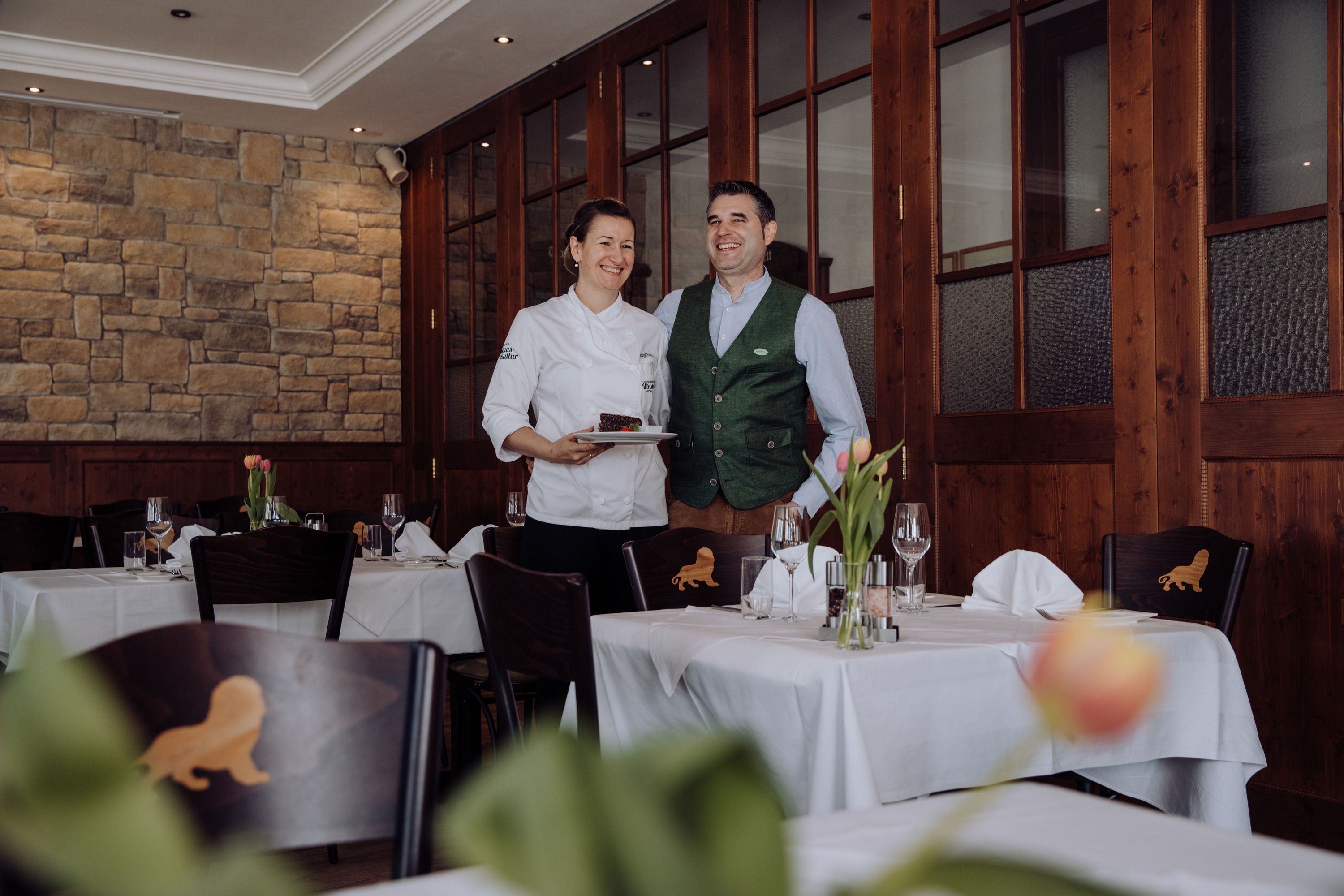 A female chef and a man in traditional dress stand smiling in an elegant restaurant.