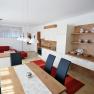 Modern living and dining area with open kitchen, red sofa, wooden table and white hanging lamps.