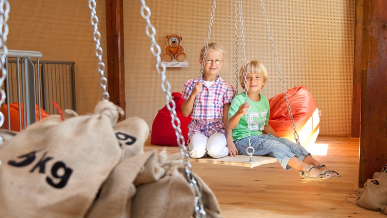 Two children sit on a swing in a play area with sandbags and beanbags.