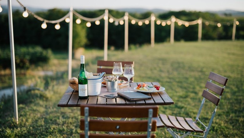 An outdoor table with wine and snacks, surrounded by fairy lights and a green landscape.