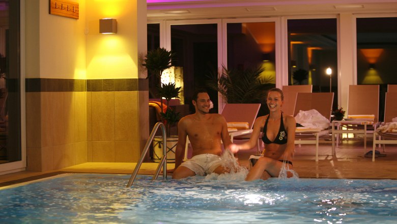 A man and a woman sit at the edge of an illuminated indoor pool and laugh. Sun loungers and plants can be seen in the background.