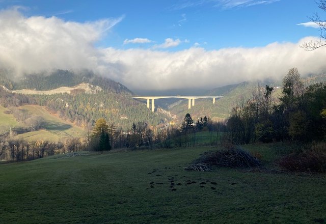 Landscape with bridge over a valley, surrounded by wooded hills and clouds.