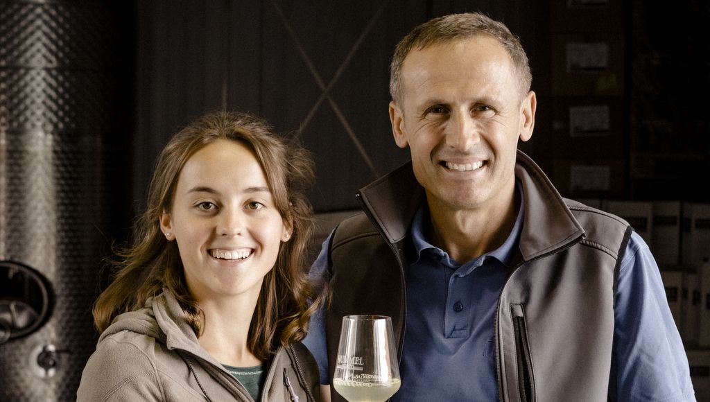 A man and a woman smile at the camera, the man holds a wine glass. They are standing in a wine cellar.