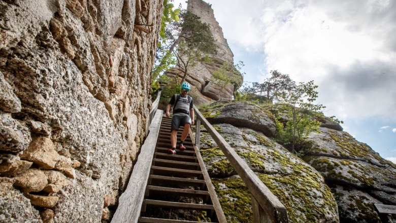 A man in a helmet walks down a wooden staircase, next to a stone castle ruin and rocks.