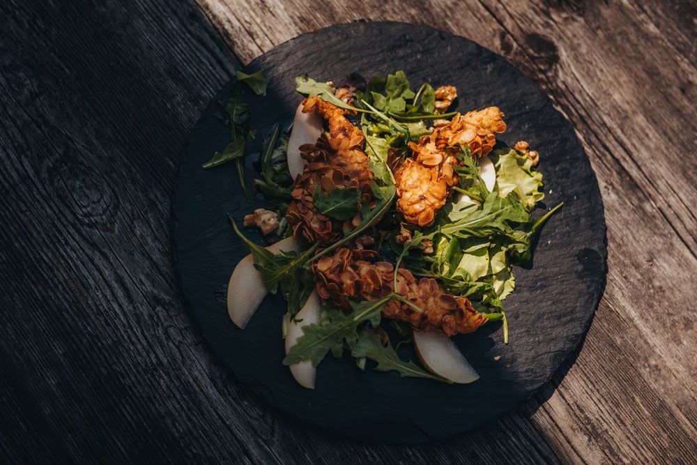 Chicken fillet with almond crust on salad with pear slices on a black plate.