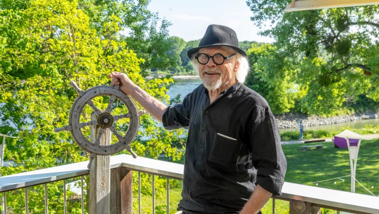 A man with a hat and glasses is standing on a balcony holding a steering wheel. Trees and a river (the Danube) can be seen in the background.