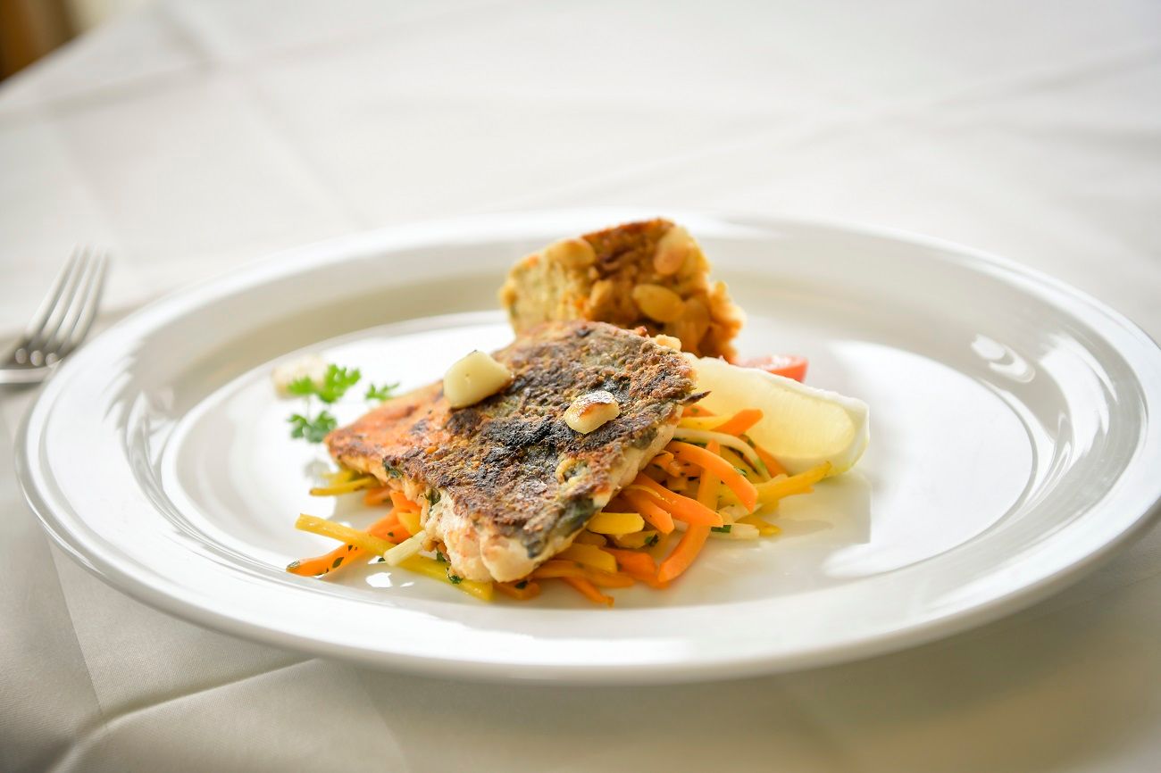 A plate with fried fish fillet on vegetable strips and a side dish on a white tablecloth.
