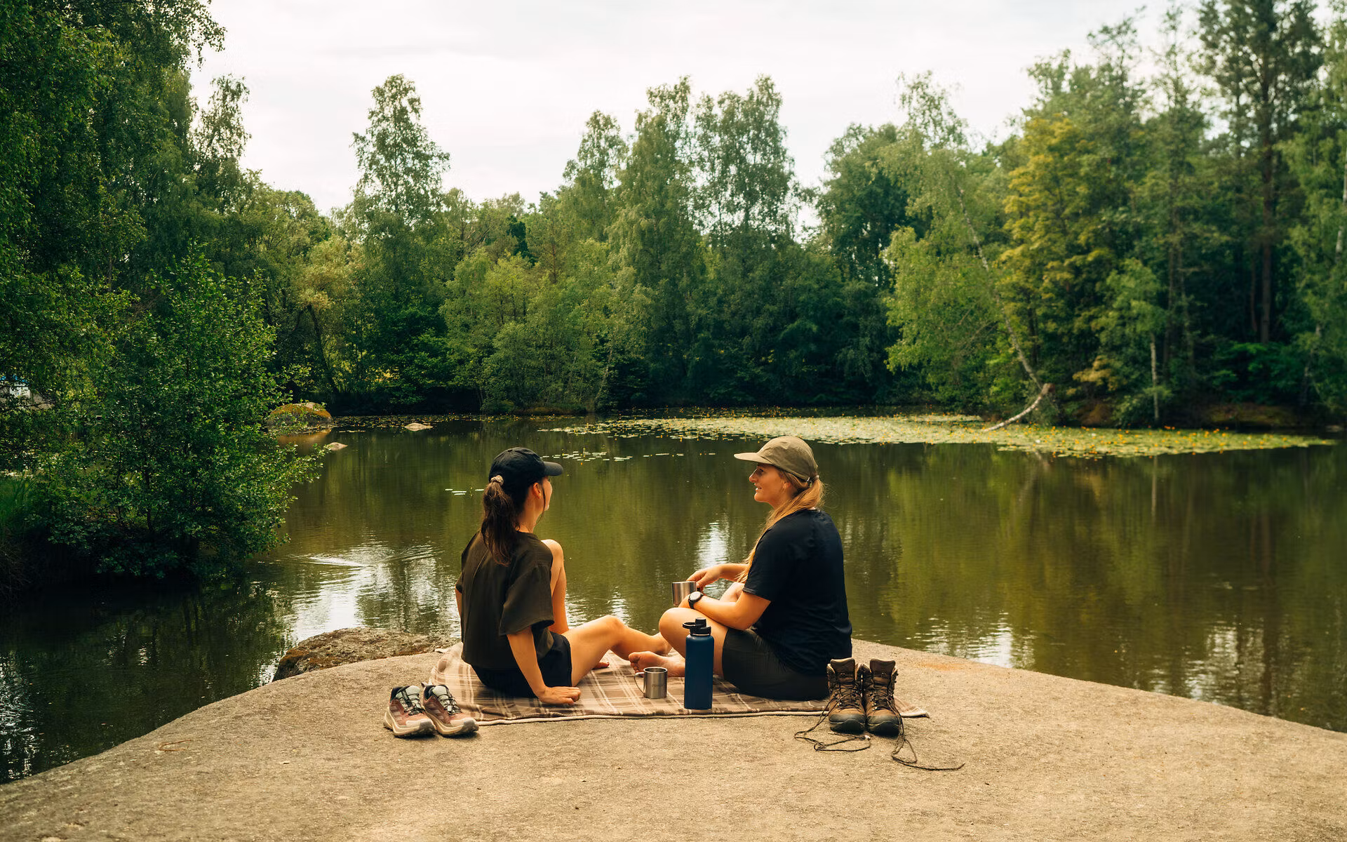 Zwei Frauen sitzen auf einem Stein in der Blockheide, umgeben von Wald und Wasser und machen eine Pause vom Wandern