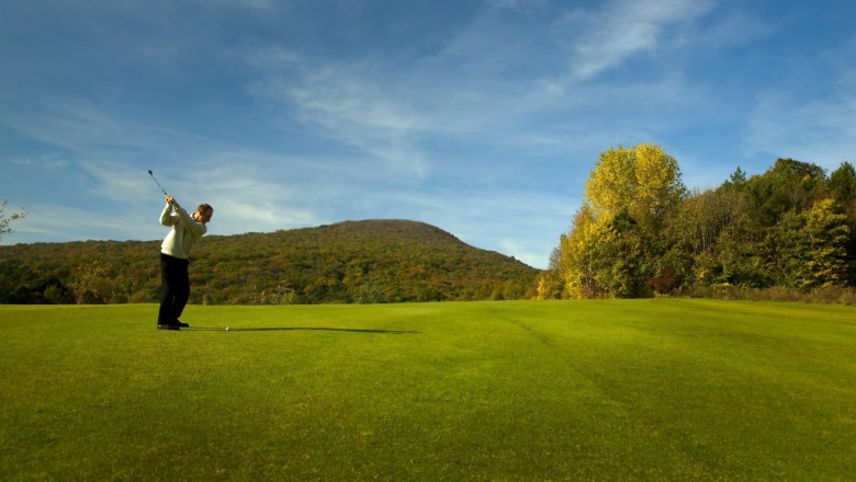 Golfer hits ball on a green golf course with wooded hills in the background.