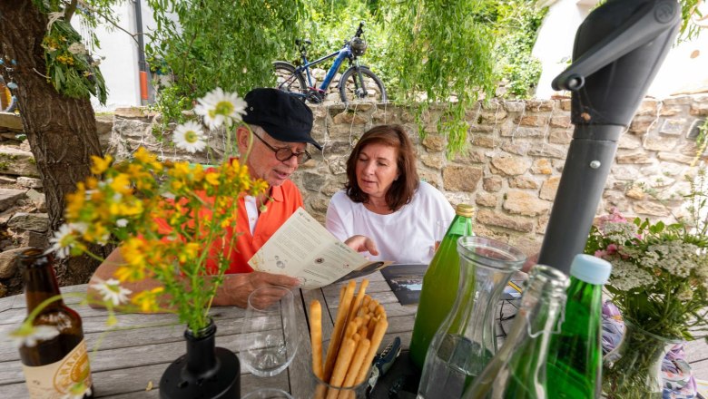 An elderly couple sits at an outdoor table, surrounded by flowers and drinks, looking at a menu.