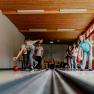 Children play skittles in a room with wooden walls and large windows.