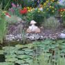 A garden pond with water lilies, colorful flowers and a frog figure on the shore.